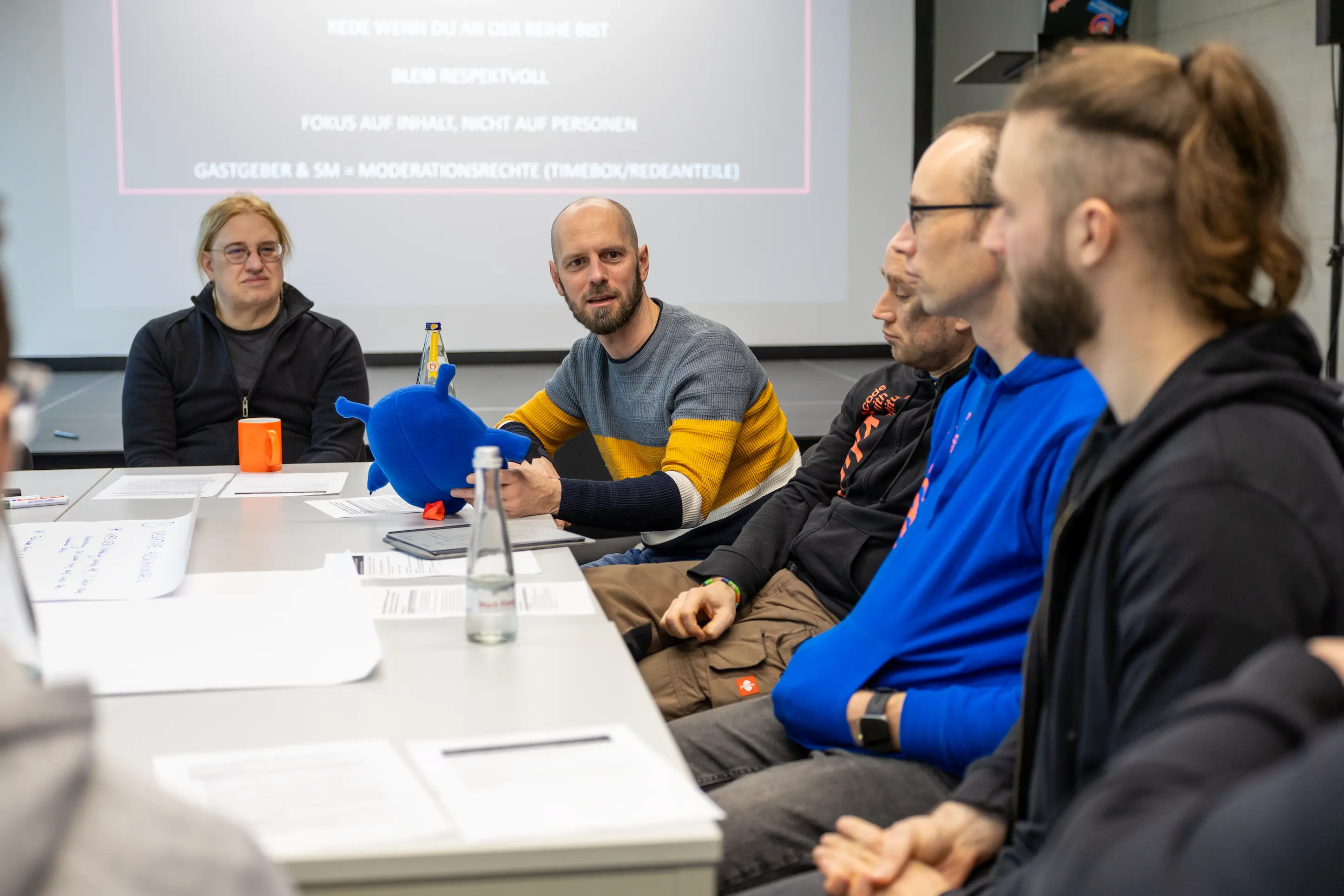 Team-Meeting mit fünf Personen am Konferenztisch, einer hält ein blaues Plüschtier.